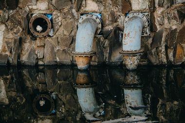Rusty industrial pipes protrude from a concrete wall, reflected in dark water below.