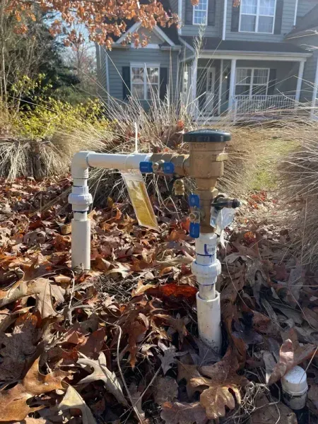 Outdoor water meter with white piping and blue valve in a leaf-covered yard in front of a house
