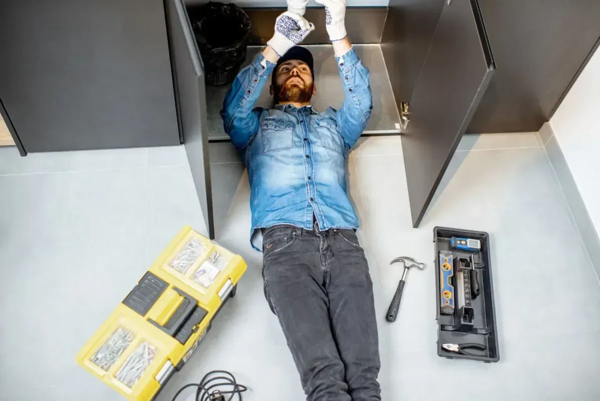 Technician lying on the floor under a cabinet, using tools and a drill for repair work.