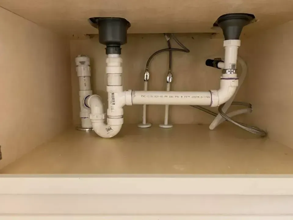 Under-sink plumbing with white PVC pipes, drain traps, and water supply lines inside a cabinet