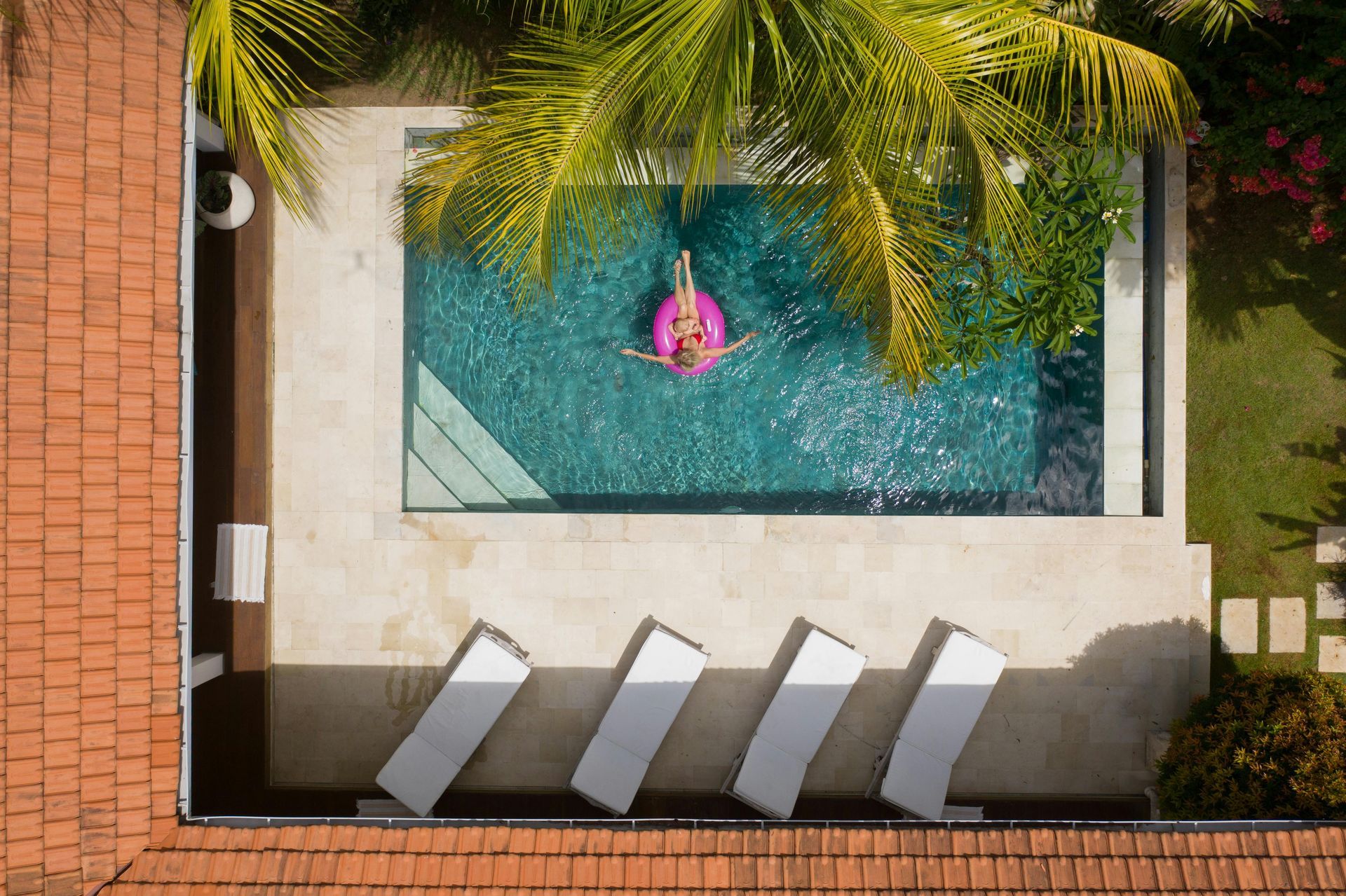 Person on pink inflatable in a rectangular pool, surrounded by white concrete, red tile roof, and palm trees.