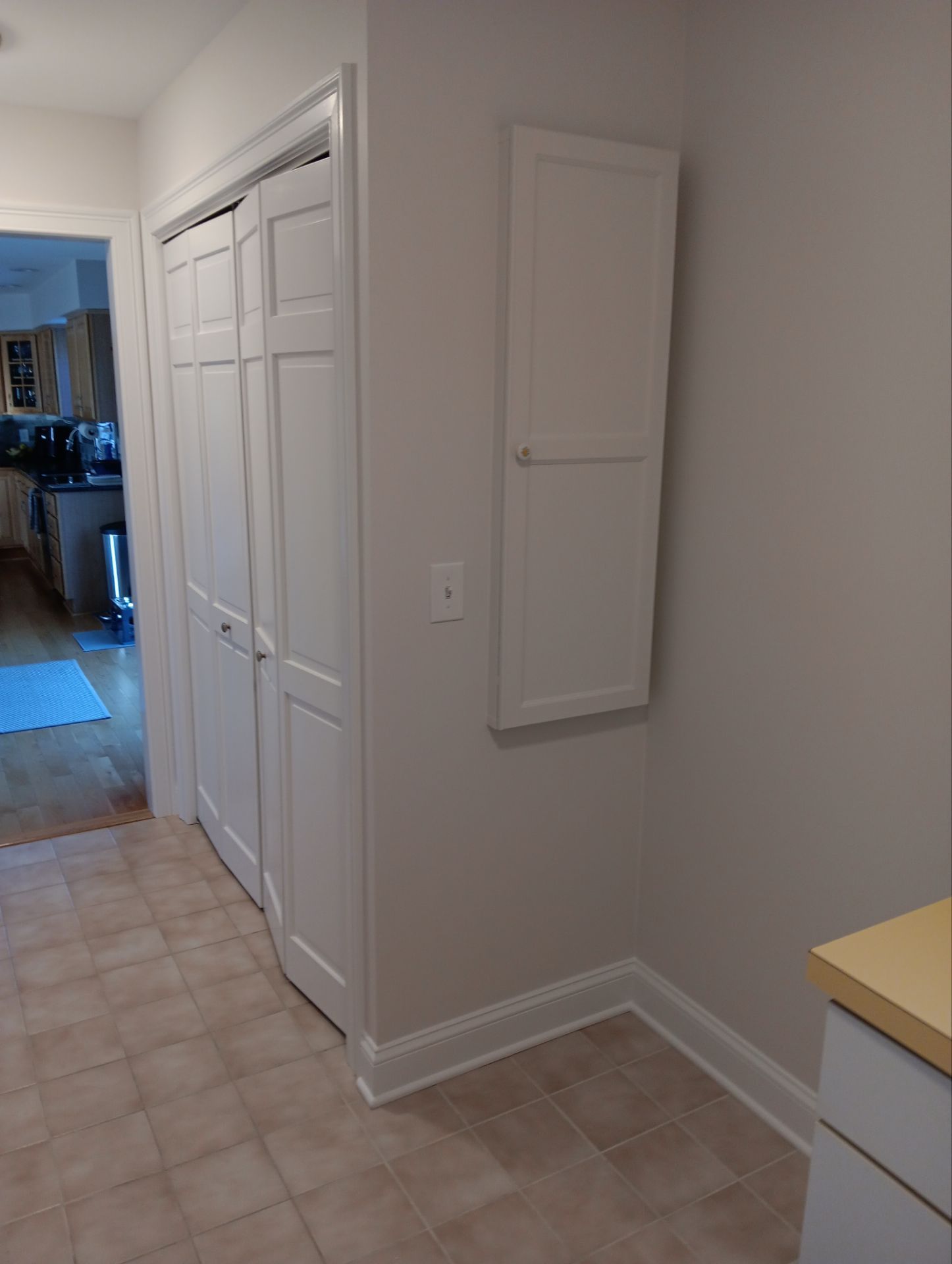 A hallway with white folding closet doors, a small white wall-mounted cabinet, and light-colored tile flooring.