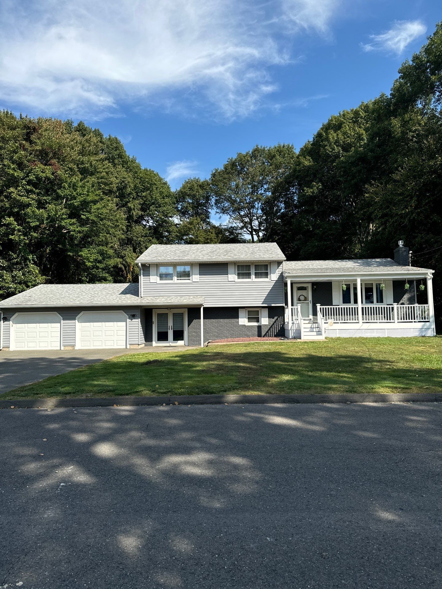 A gray two-story house with a two-car garage, white trim, and a front porch, set against a backdrop of lush green trees.