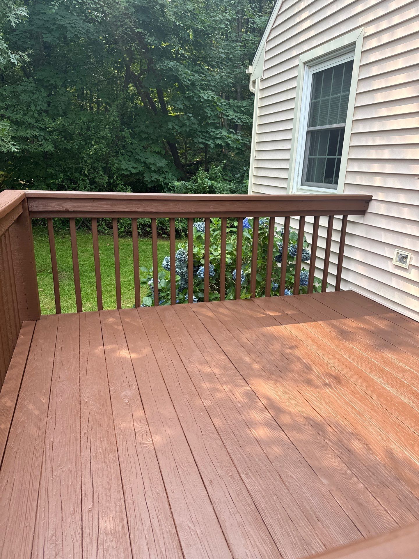 A wooden deck with a brown railing, extending from the side of a light-colored house toward a background of green trees.