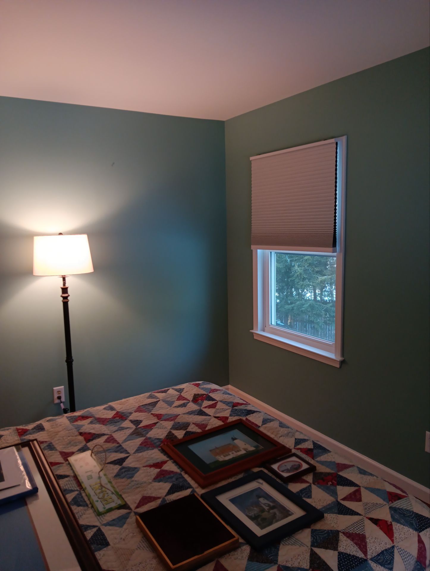 A corner of a bedroom with muted green walls, a floor lamp, a window with a pleated shade, and framed art on a quilt bed.