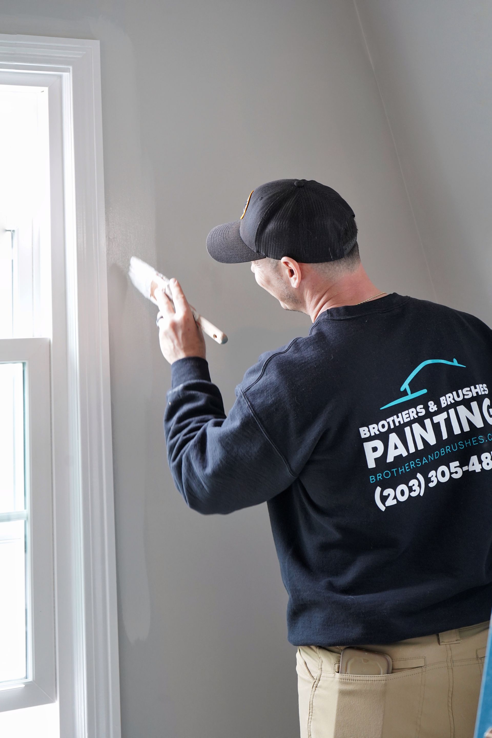 A person wearing a dark company sweatshirt and cap uses a brush to paint the wall beside a white window frame.
