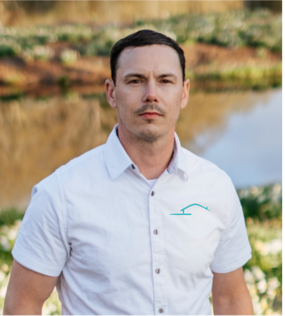 A person with short dark hair in a white collared shirt stands outdoors near a pond.
