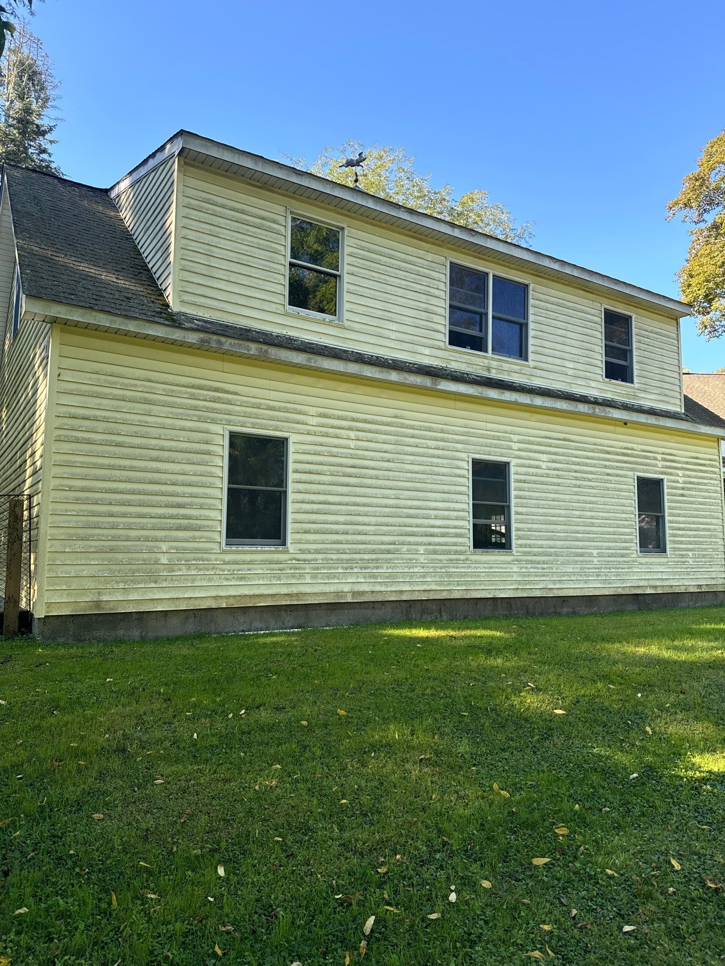 A two-story light yellow house with light-colored siding and several windows stands on a grassy lawn under a clear sky.