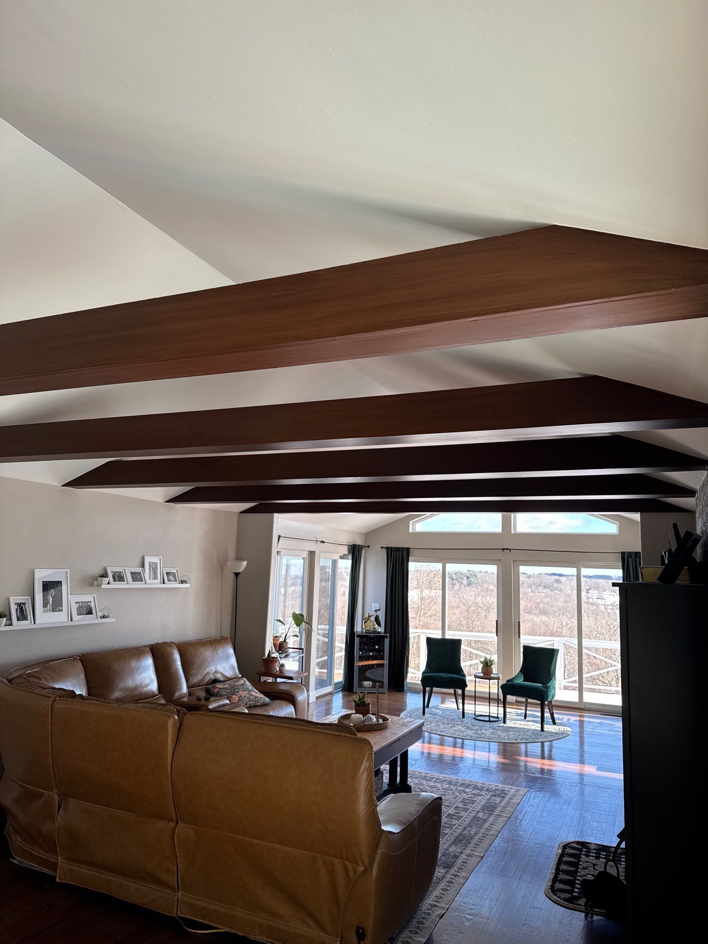 A living room featuring dark wooden ceiling beams, a large brown leather sofa, and sliding glass doors leading to a deck.