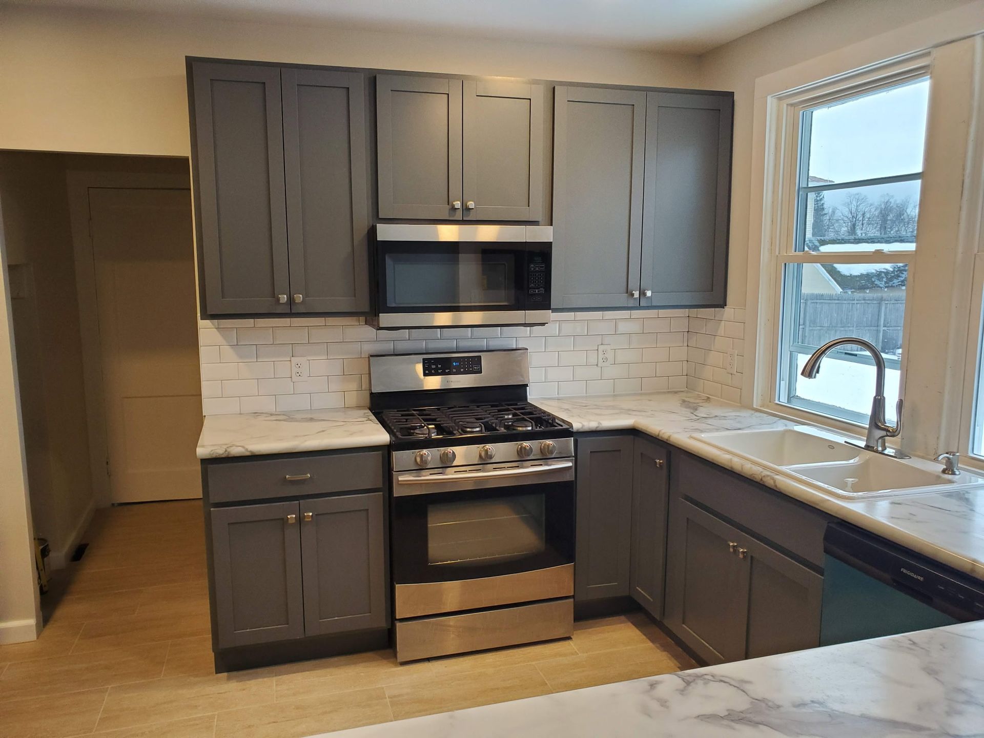 Modern kitchen with gray cabinets, stainless stove and microwave, white tile backsplash, and window over sink