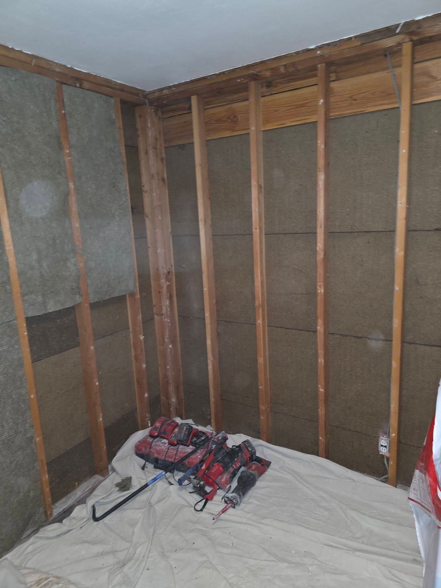 Partially gutted room with exposed wall studs, insulation, and tools on a tarp-covered floor