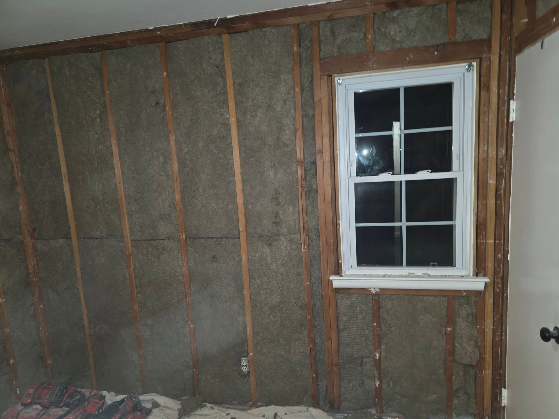 Interior wall with exposed insulation and wooden studs beside a small window and door frame