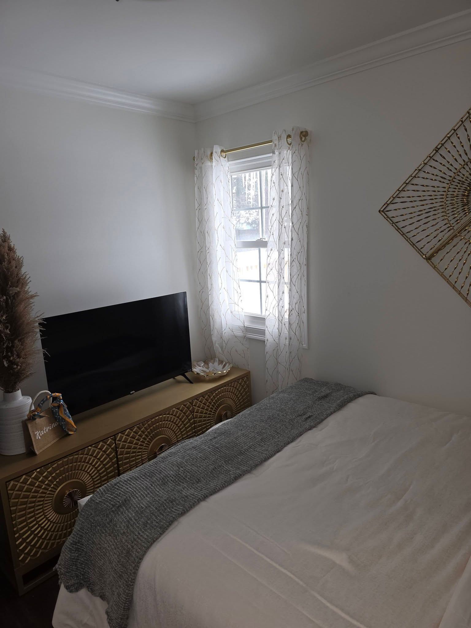 Bedroom with a bed, TV on a woven dresser, and a window with sheer curtains.