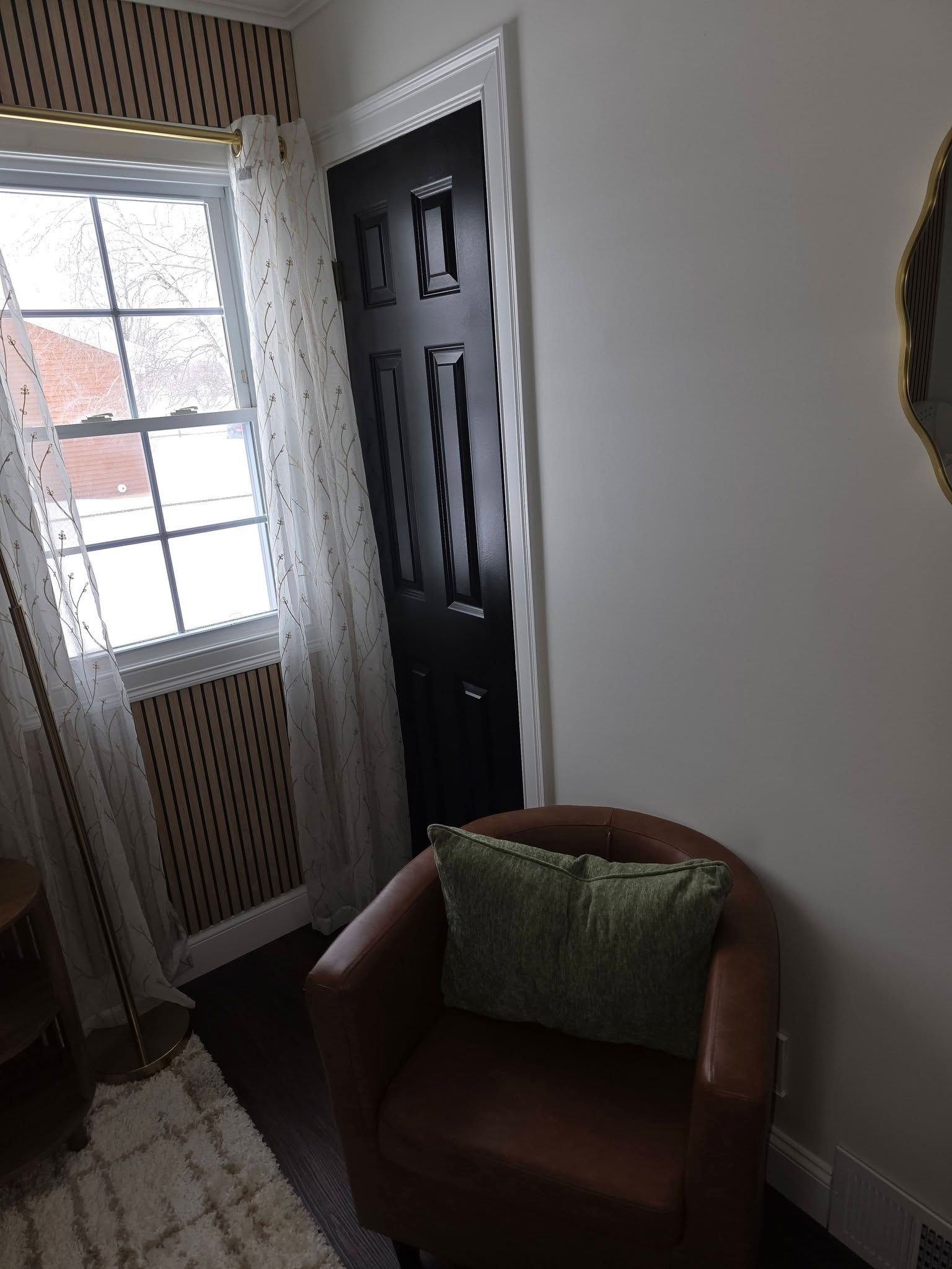 Cozy corner with a brown armchair and green pillow beside a white wall and window with sheer curtains.