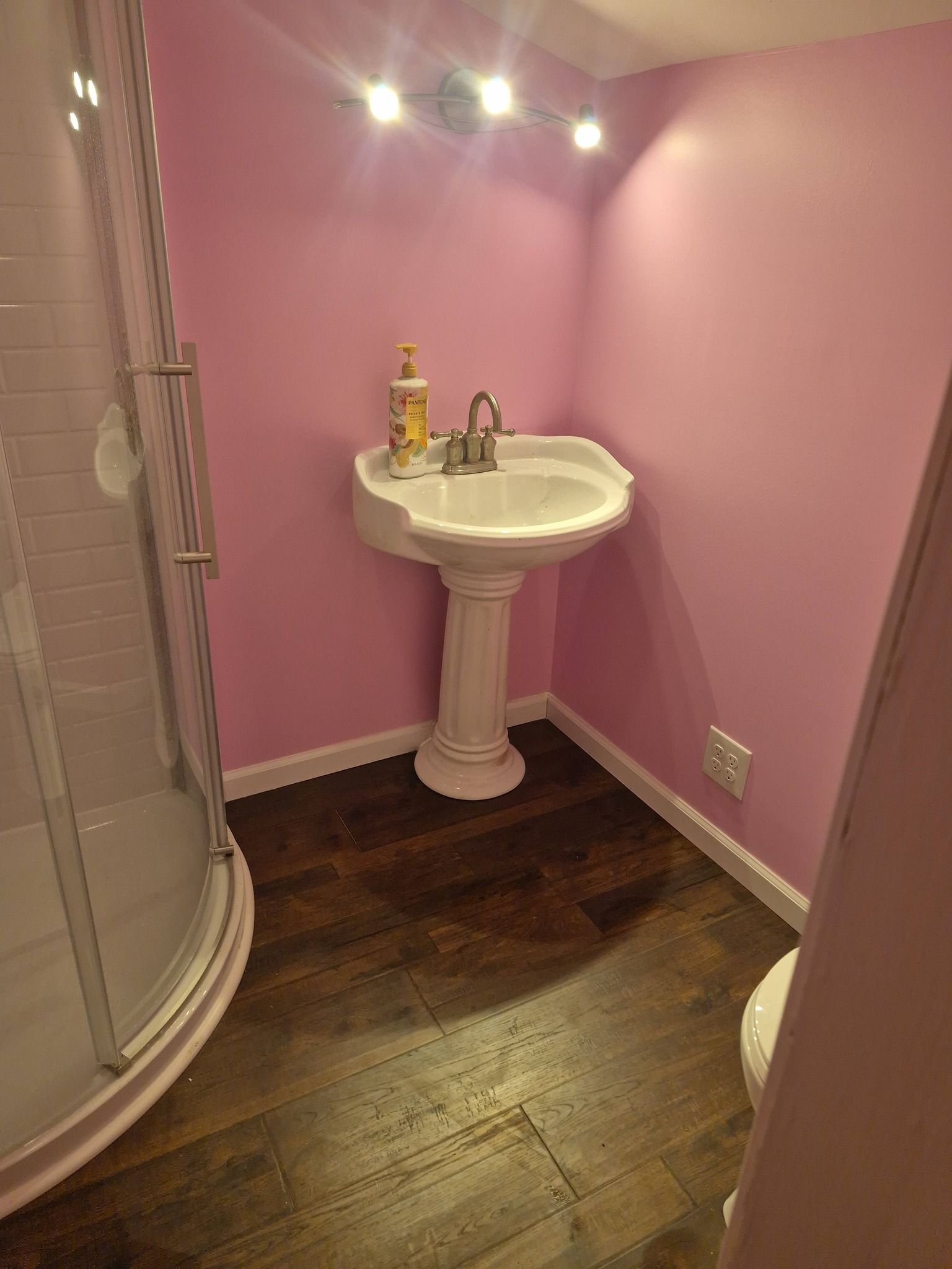 Small pink bathroom with pedestal sink, wall lights, dark wood floor, and shower on the left.