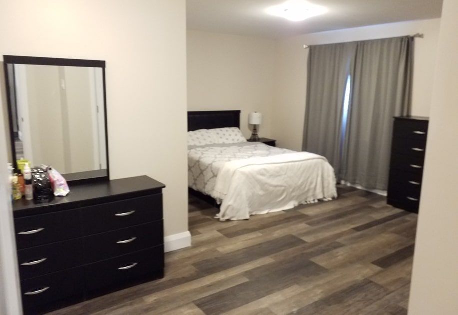 Bedroom with bed, dresser, mirror, and gray curtains on wood-look flooring.