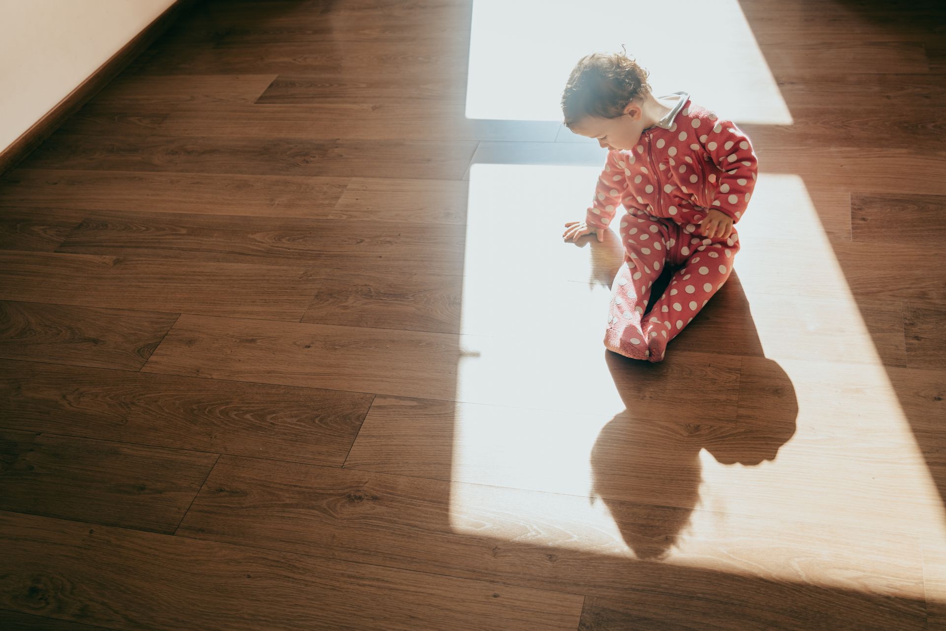 Child in a red patterned outfit crouches in a sunlit doorway on a wooden floor.