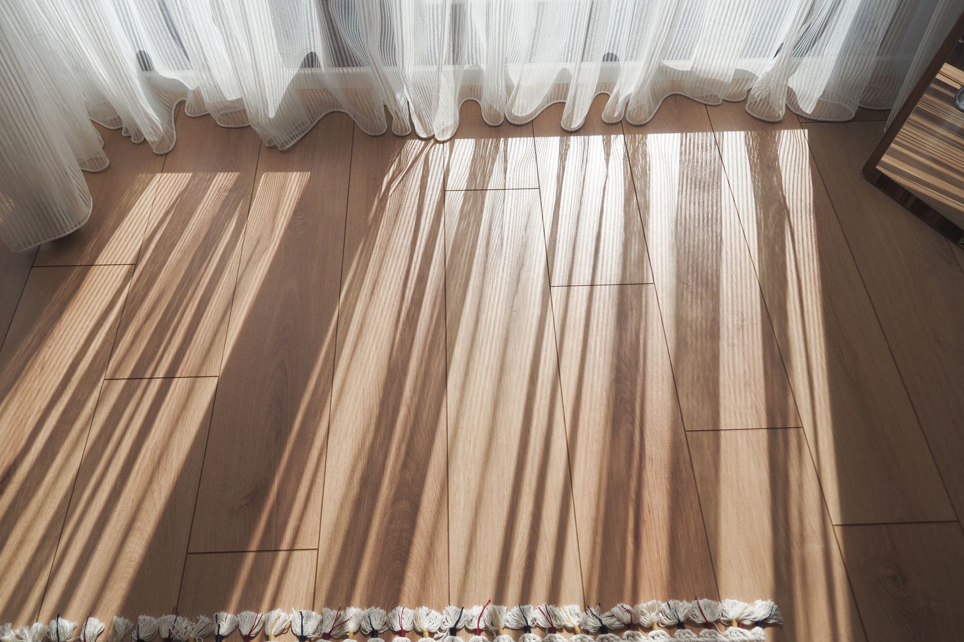 Sunlit wooden floor with long striped shadows from sheer curtains and soft drapes.