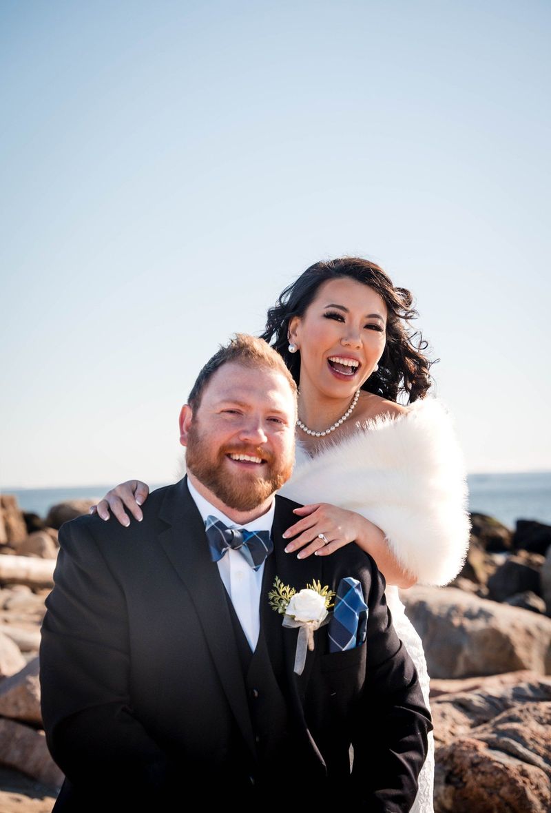 Amanda leans over Charles’s shoulders as they laugh beside the water