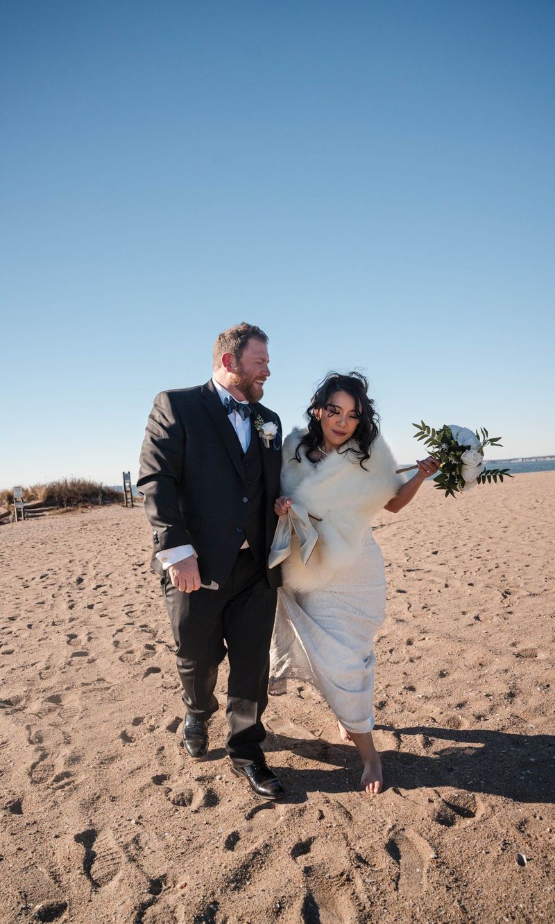 The couple walk arm in arm toward the camera along the beach