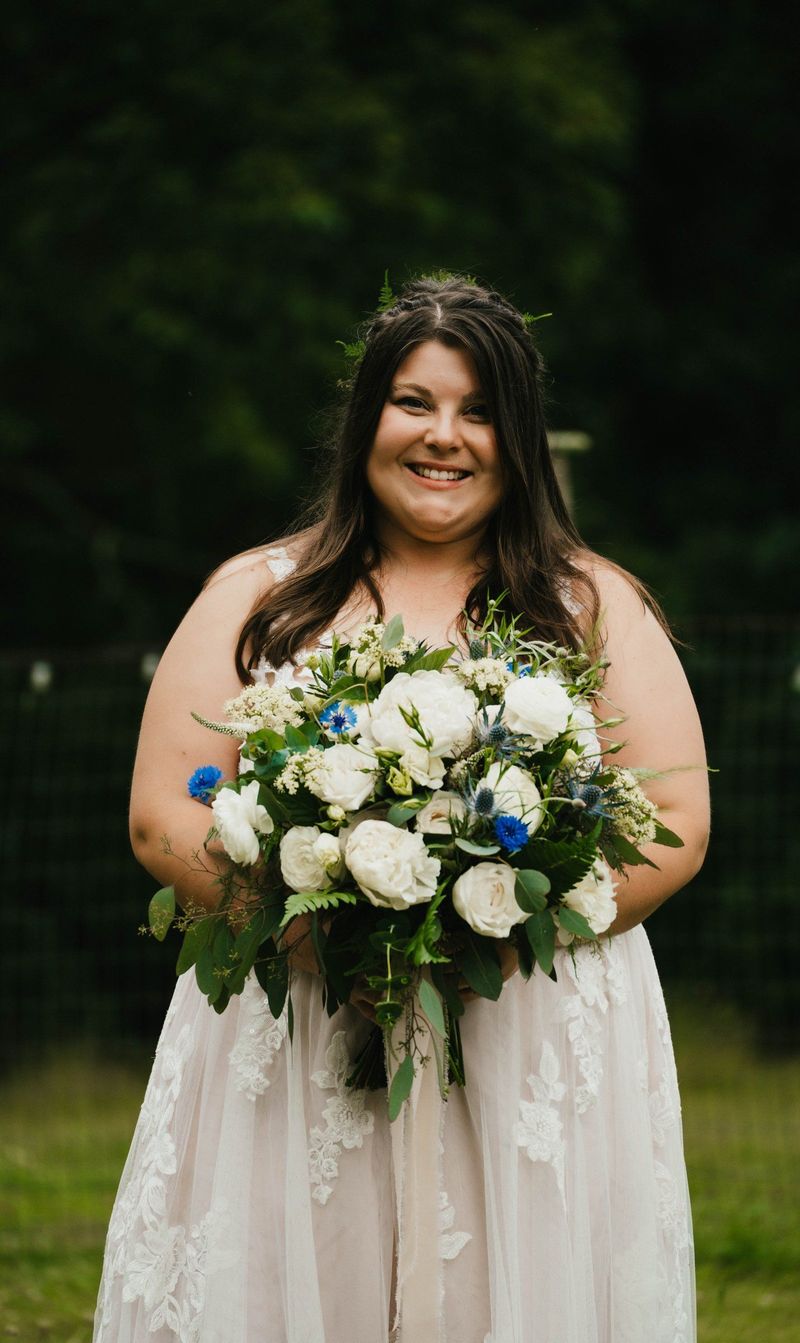 Ashley smiles with her white and blue bouquet against a green backdrop
