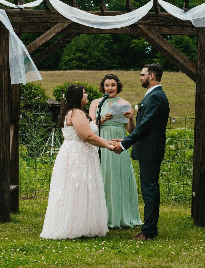 Ashley and Anthony stand beneath a wooden ceremony arbor with white draping