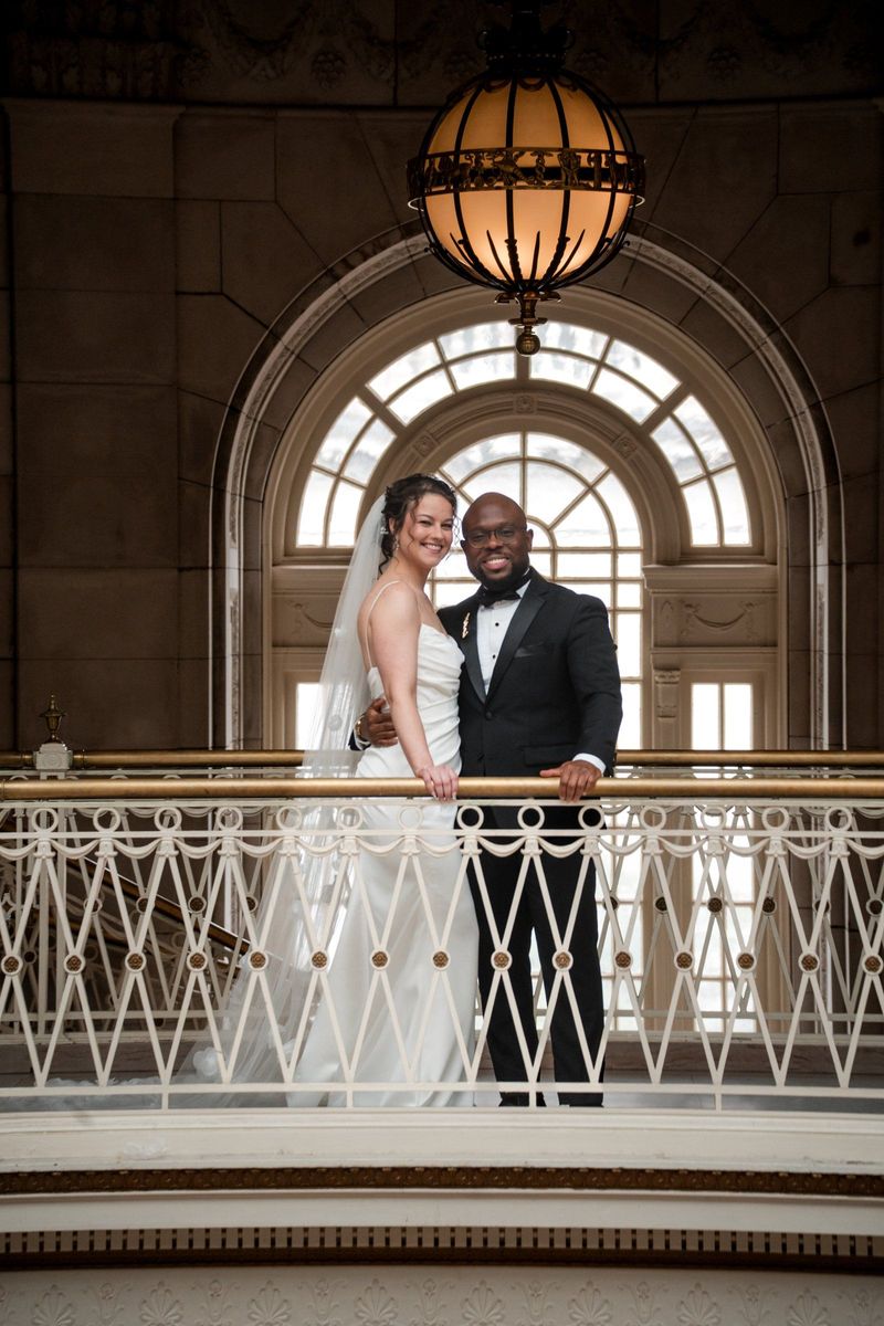 Christina and Tajay pose together behind the balcony railing with an arched window behind them