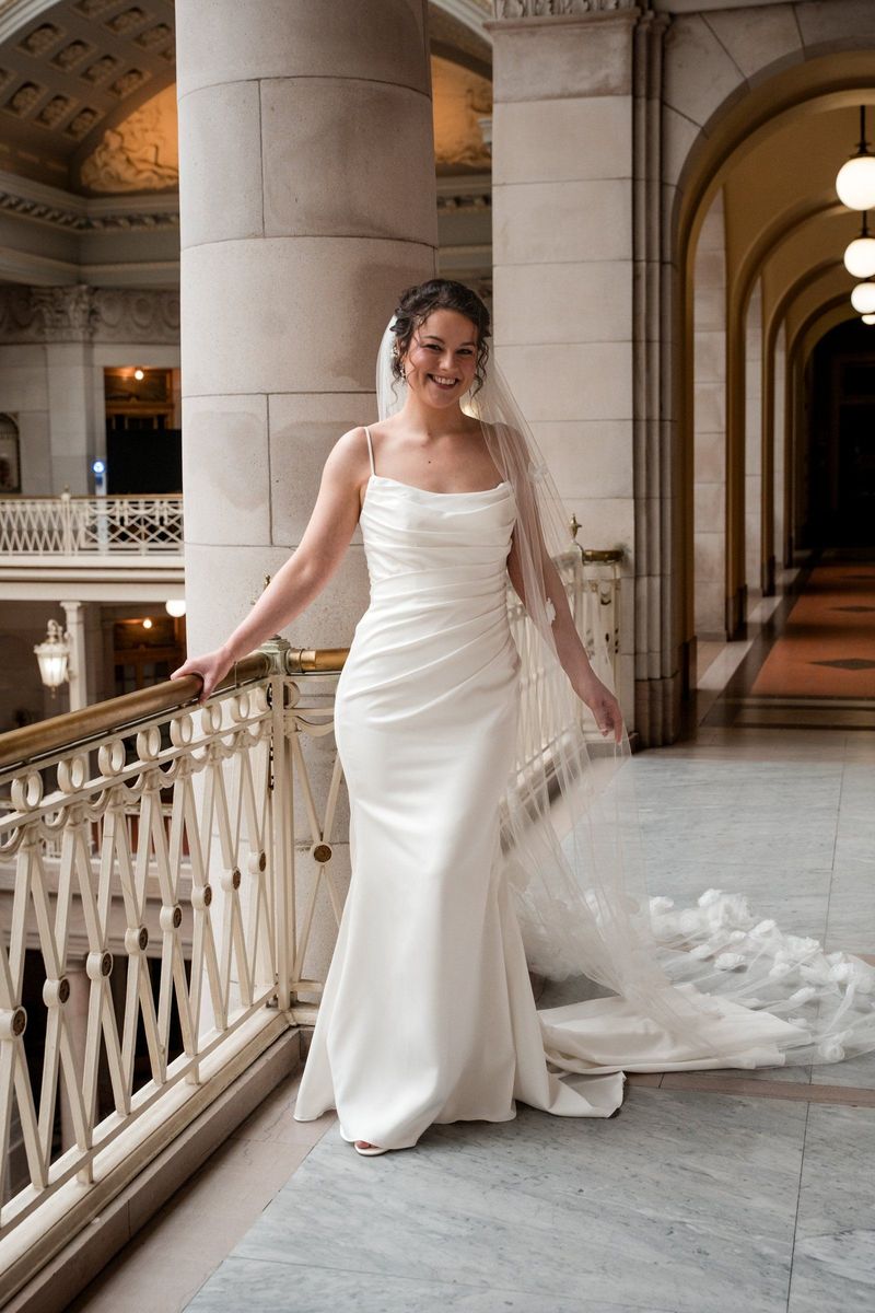Christina smiles in a full-length bridal portrait with her gown and veil beside the balcony railing