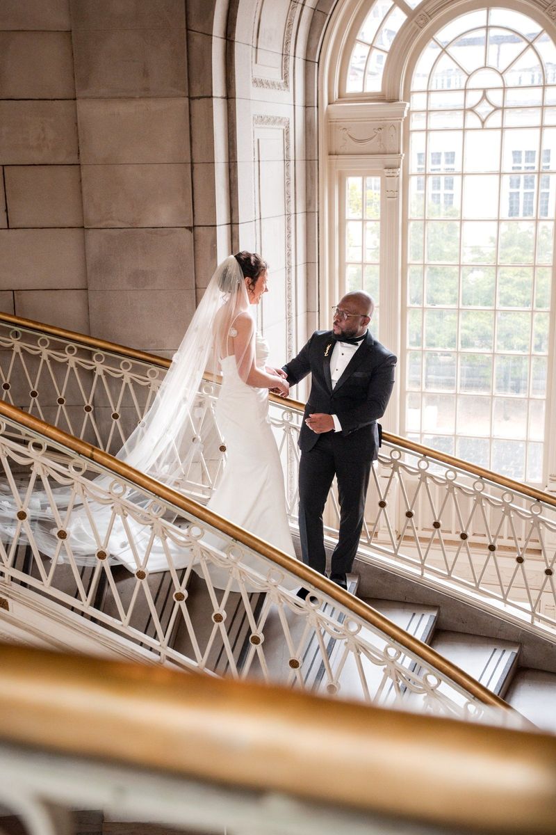 Christina holds Tajay’s hand beside the tall arched staircase window