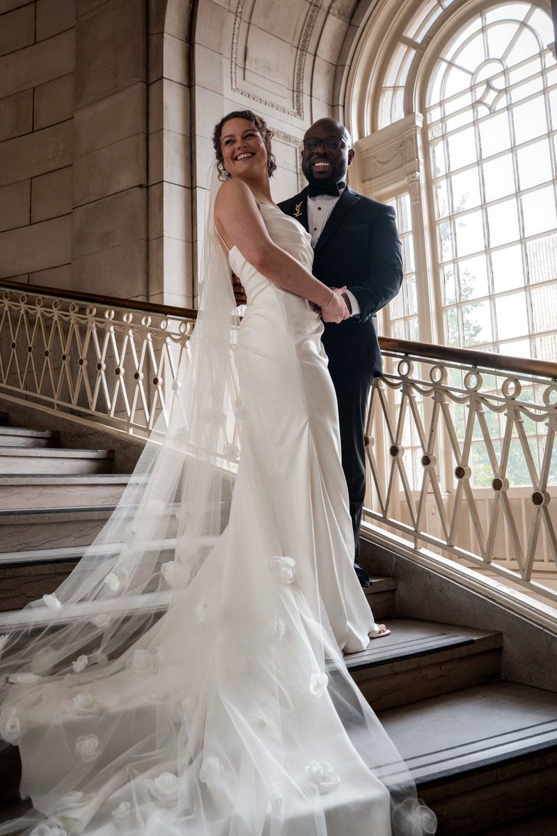 Christina and Tajay stand together on the staircase with her lace veil spread along the steps