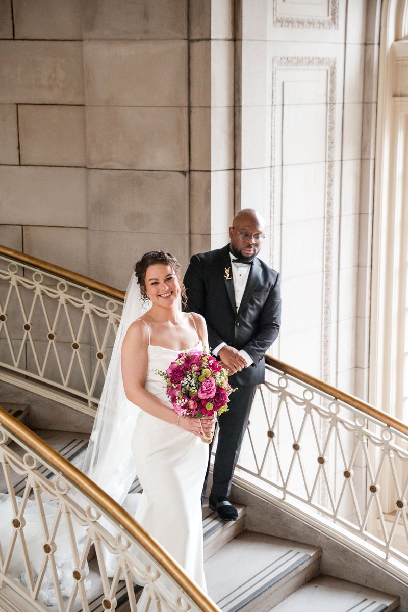 Christina holds her pink bouquet with Tajay behind her on the staircase