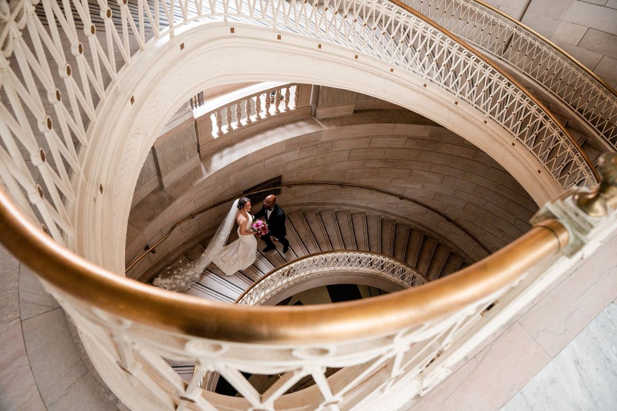 An overhead view follows the curved brass stair rail toward Christina and Tajay below