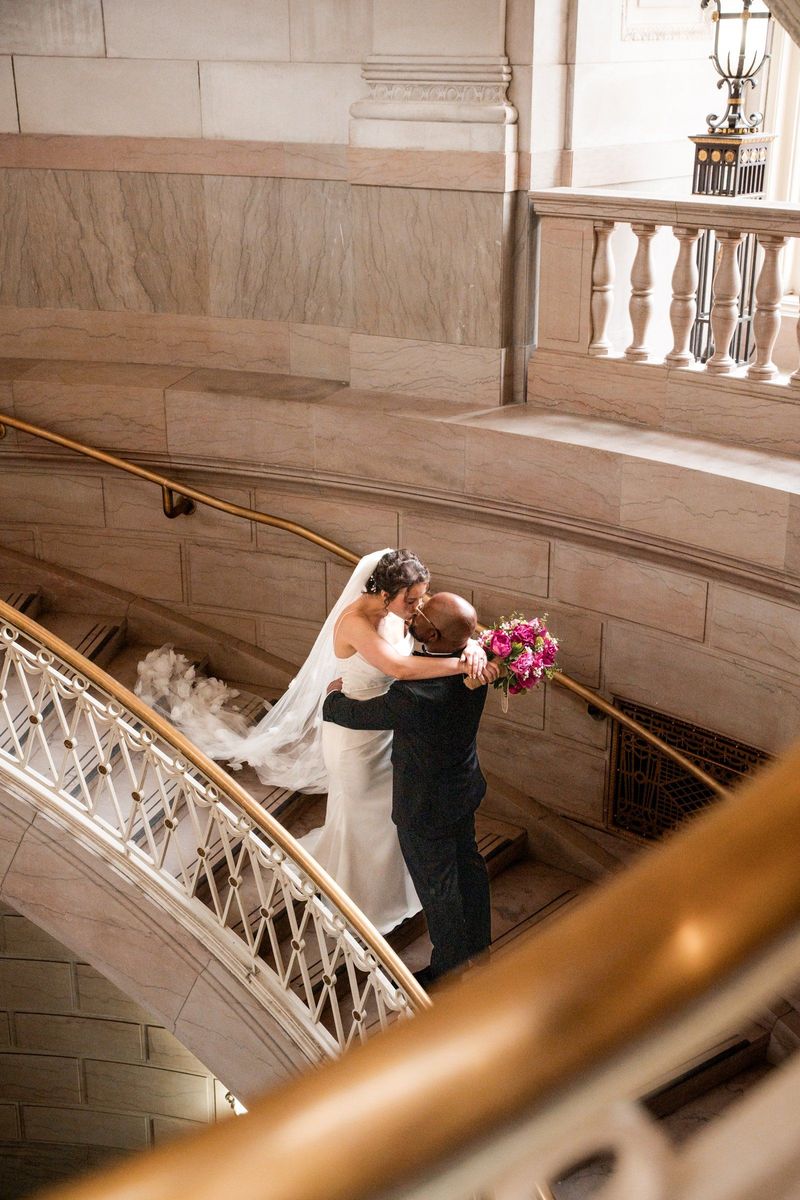 Christina and Tajay embrace on the staircase, seen from above through the brass rail
