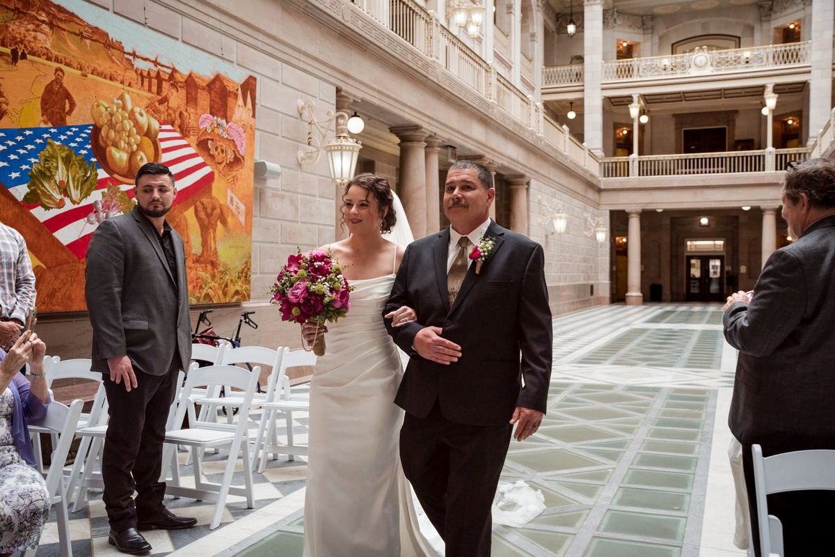 Christina walks down the atrium aisle carrying her bouquet beside a loved one