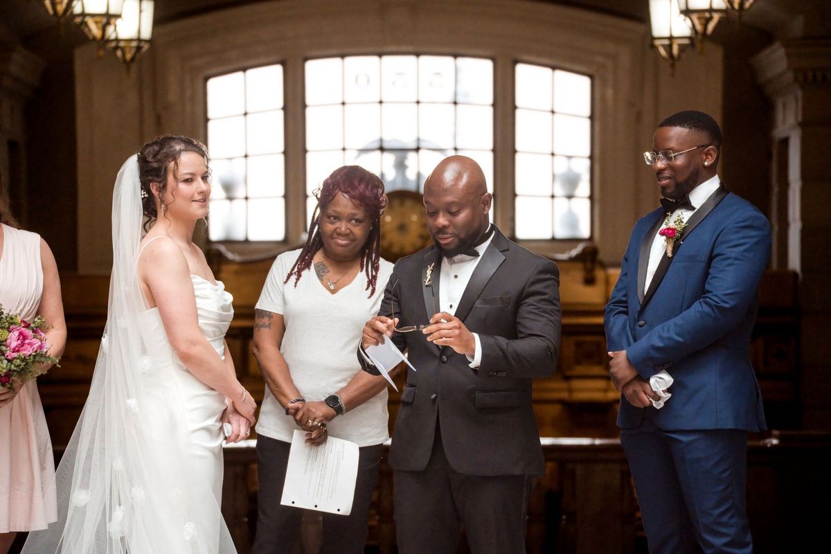 Christina and Tajay face their guests beside the officiant beneath the arched window