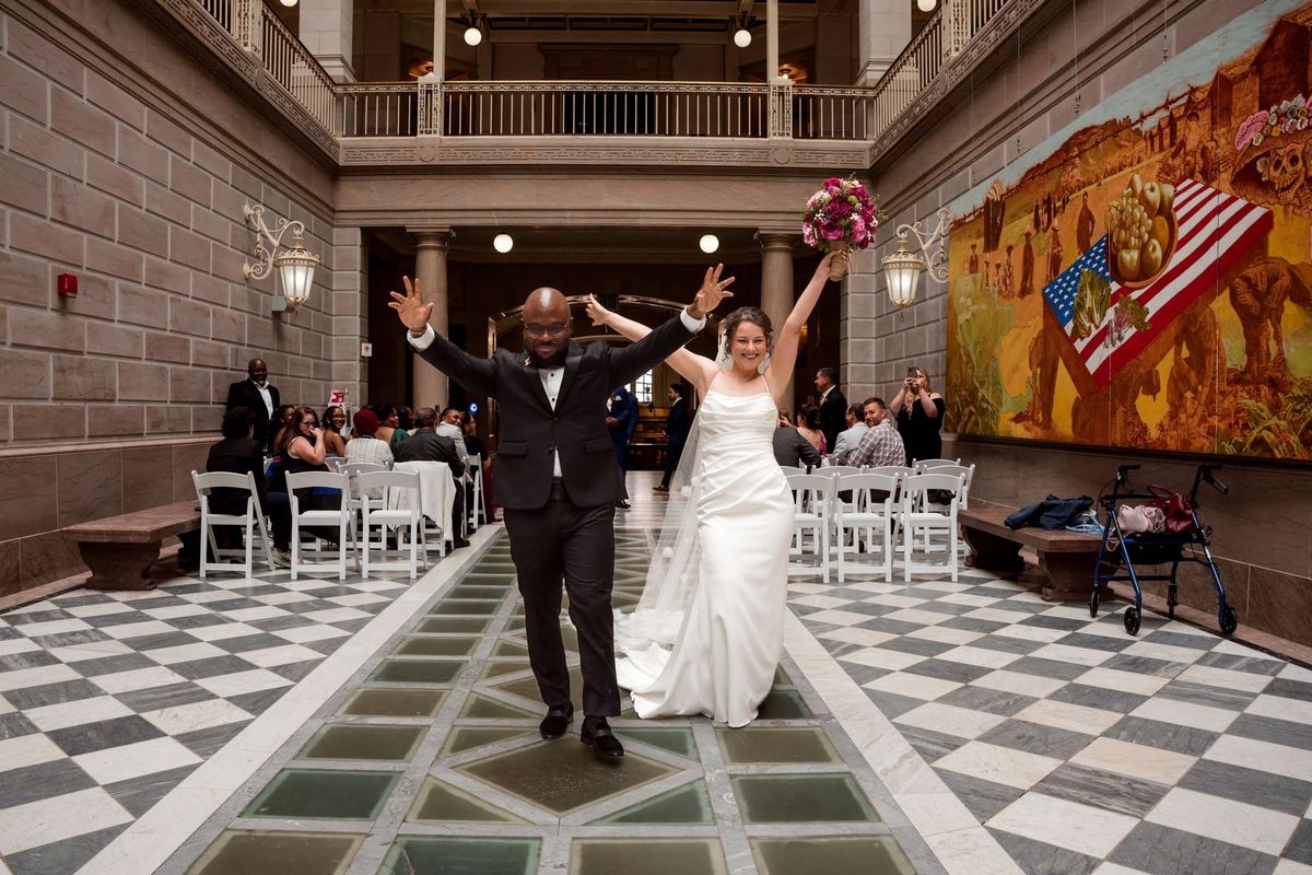 Christina and Tajay walk down the patterned atrium aisle with their arms raised