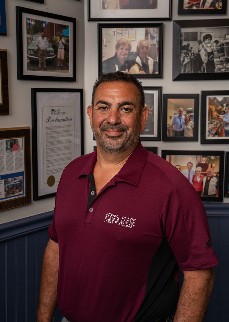Effies owner in a burgundy polo shirt standing in front of a wall of framed photographs