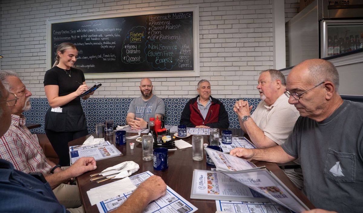 Dining room at Effies with a server standing beside a table of seated people and a board on the wall behind