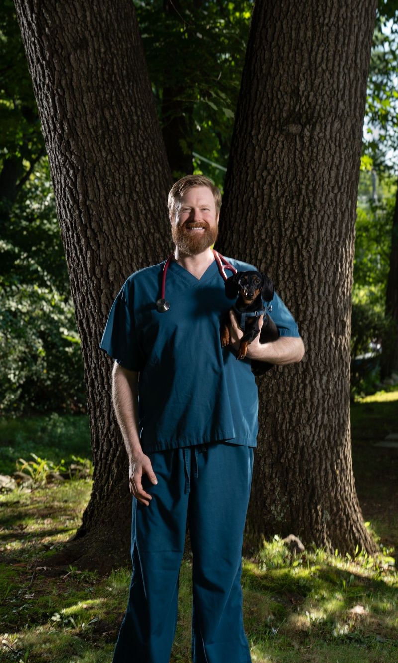Full-length Farmington Animal Hospital portrait of a man in scrubs holding a small black dog among trees