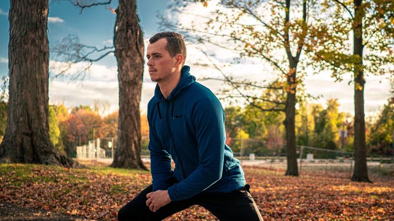 A man in a blue zip hoodie stretching in a park among autumn leaves
