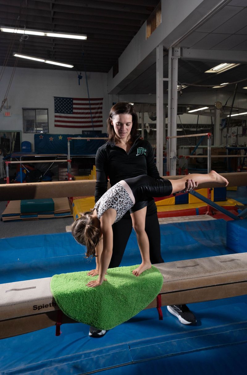 A coach in a GTC jacket spotting a young gymnast in a leopard-print leotard on the balance beam