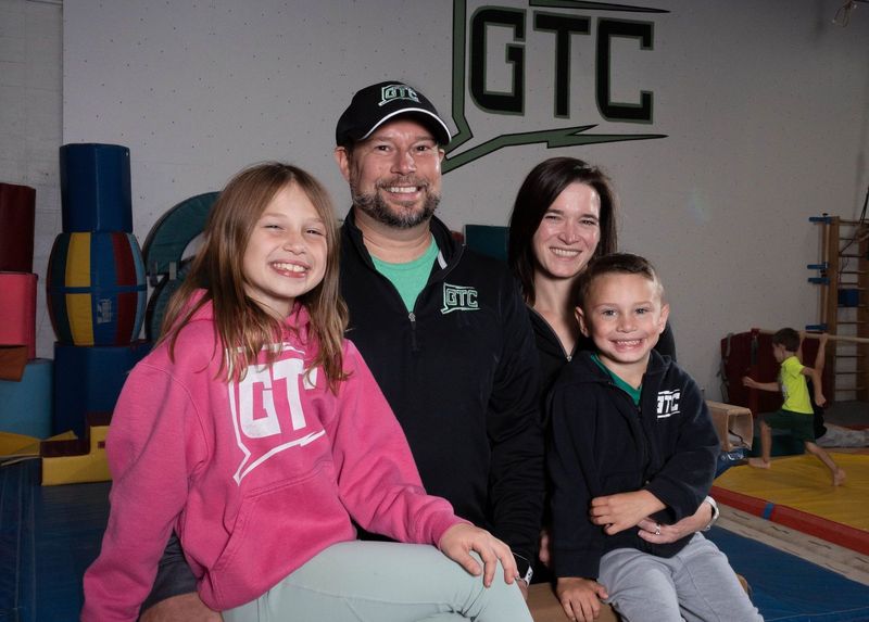 The owners of Gymnastics Training Center and two children smiling in front of the GTC logo on the gym wall
