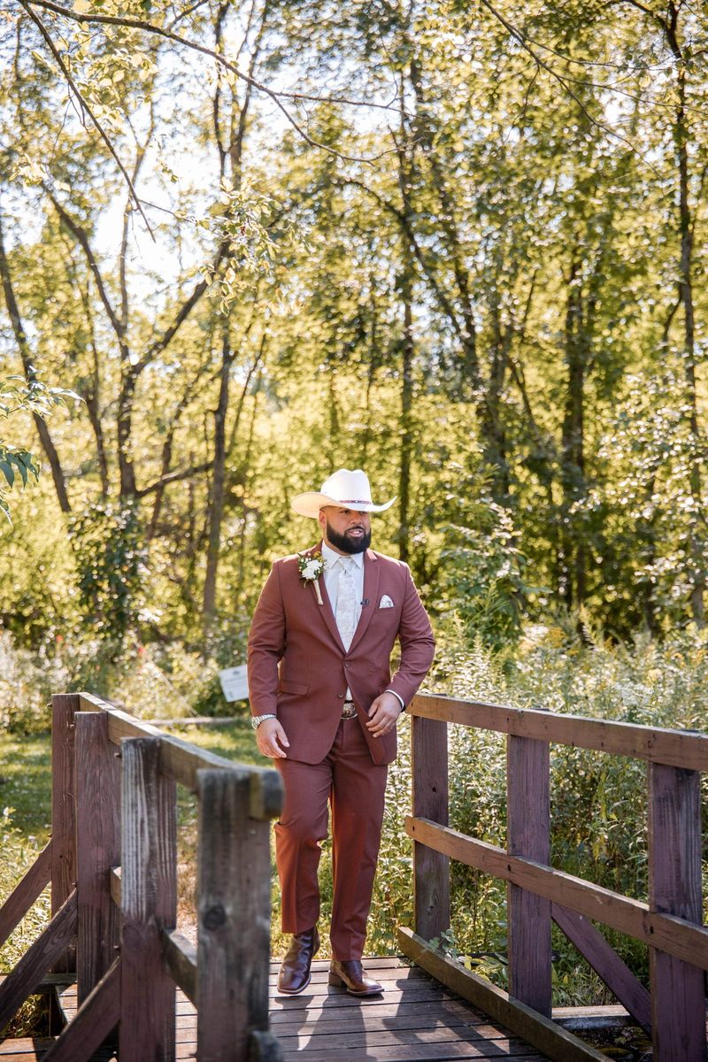 Jacob stands on a wooden bridge in a rust-colored suit and white cowboy hat