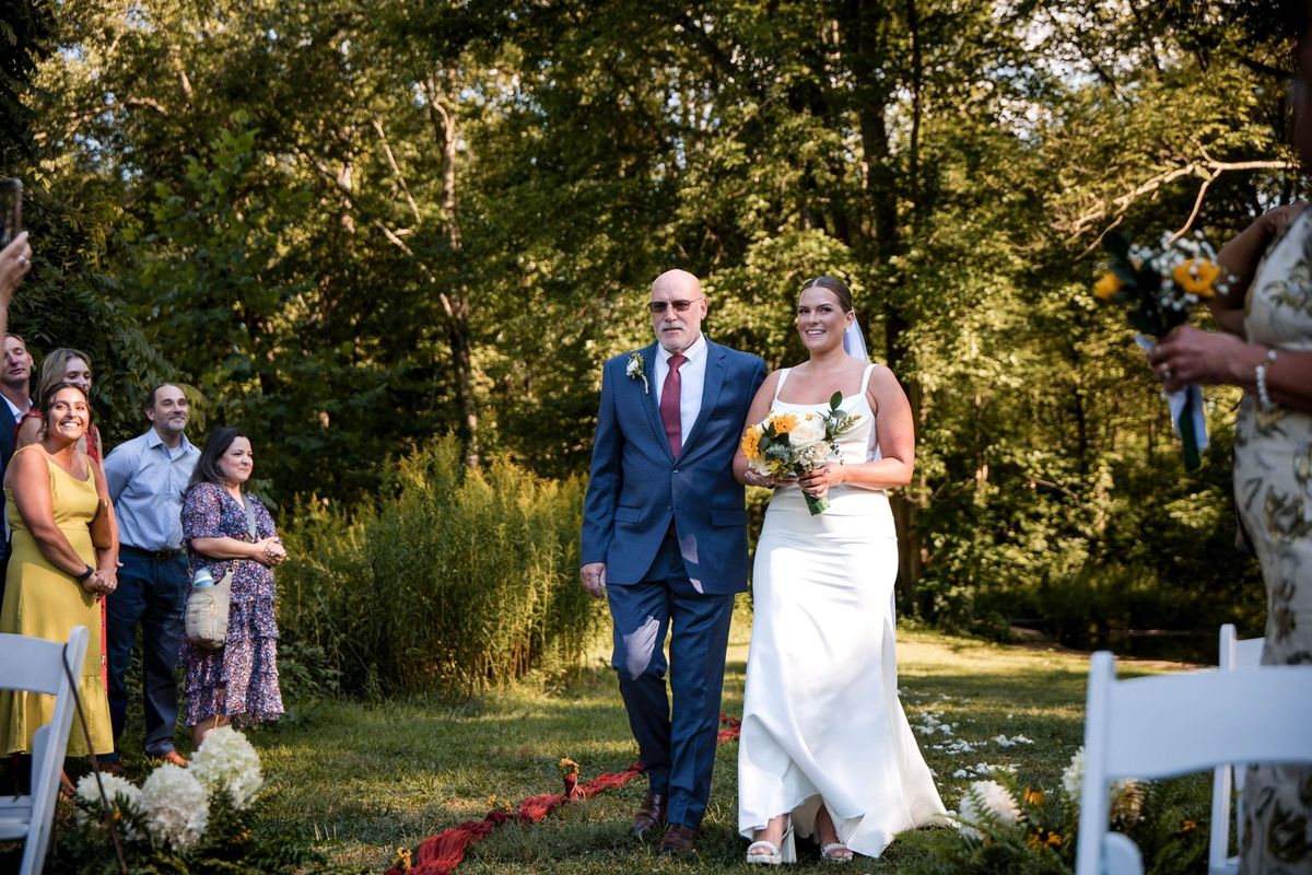 Thea walks down the outdoor aisle beside a loved one in a blue suit