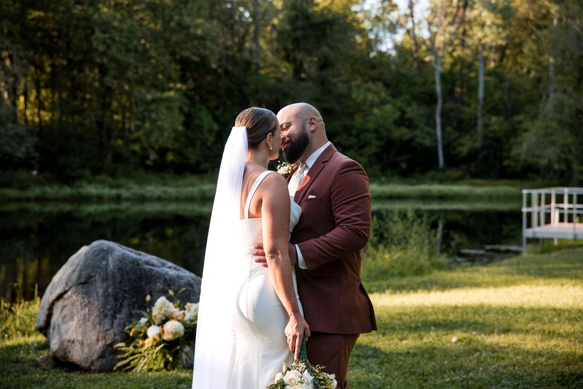 Jacob and Thea lean together beside the pond with woodland behind them