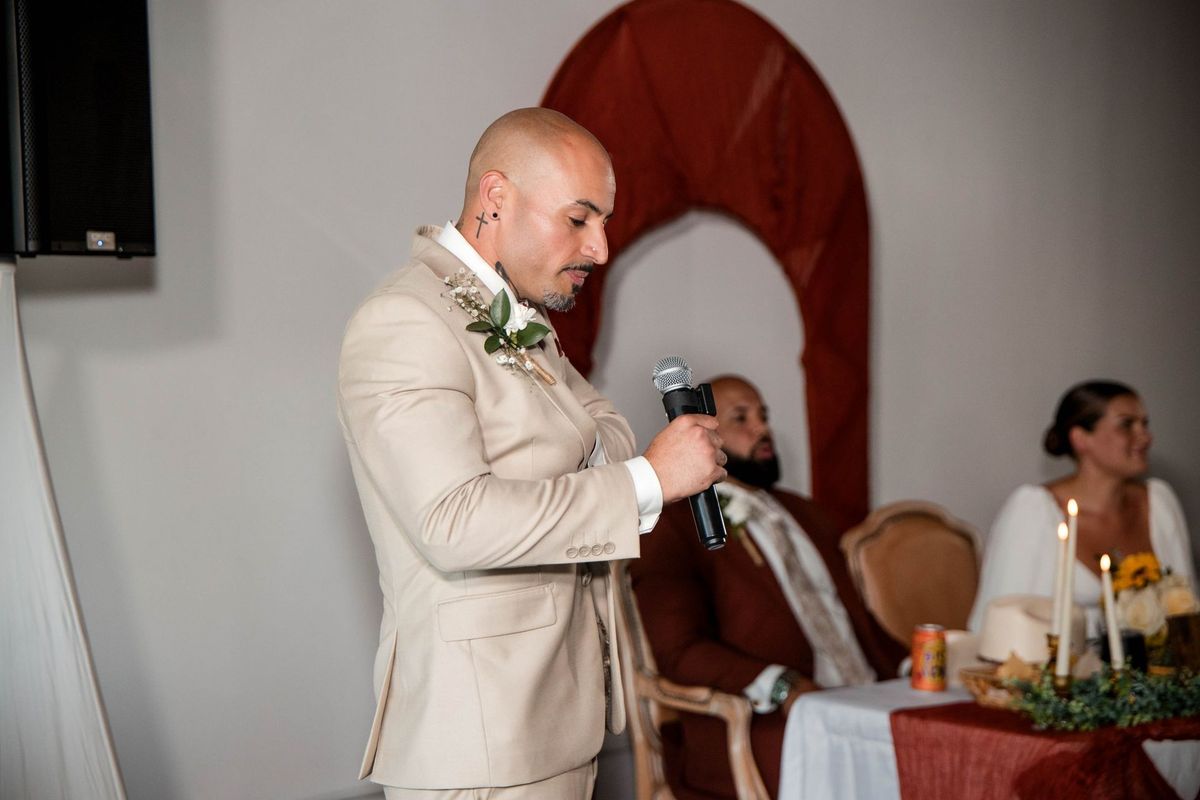 A speaker in a light suit holds a microphone beside the couple’s table