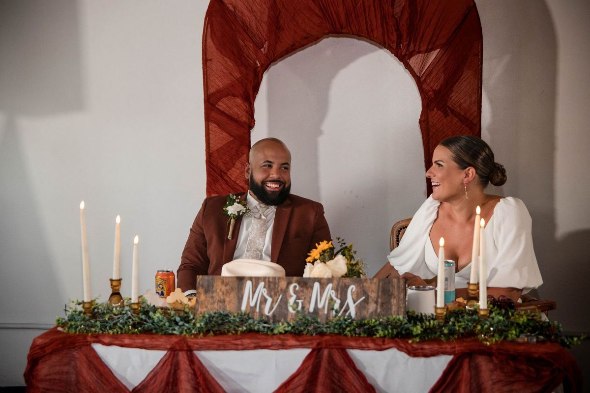 Jacob and Thea smile at one another while seated at their sweetheart table