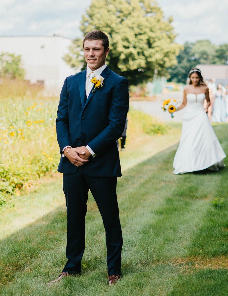 Max smiling in the foreground as Mariana approaches behind him for their first look