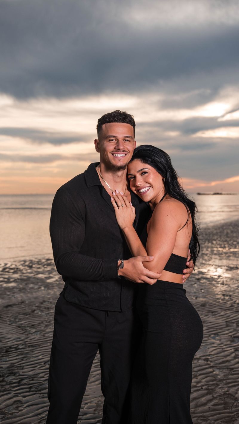 A smiling couple in black outfits embracing on a beach under a cloudy sunset sky
