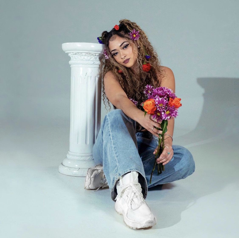 A woman with curly hair and flowers in it sitting beside a white column, holding a bouquet of orange and purple flowers